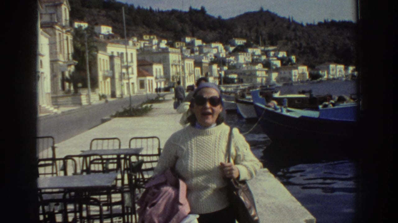 Close shot of woman in cream cable-knit sweater smiling toward camera on a Greek harbor promenade, 1976; moored boats and cafe chairs visible.