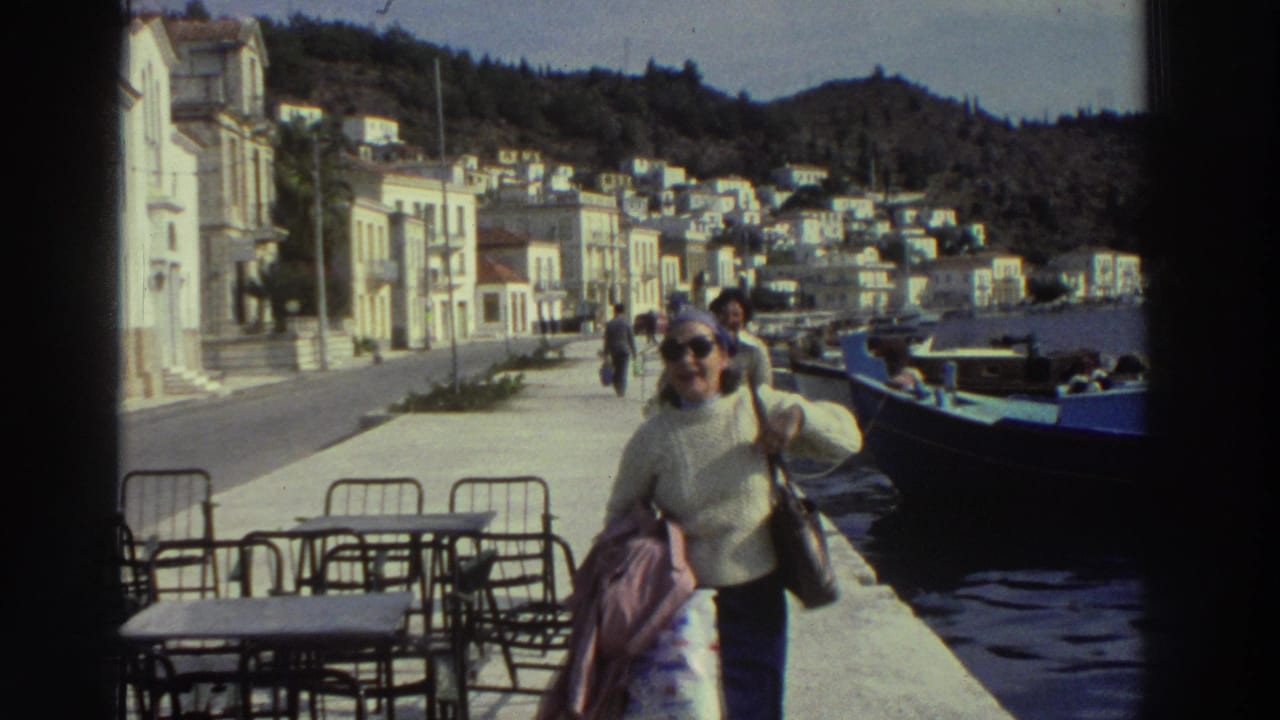 Wider shot of woman walking toward camera on a Greek harbor promenade, 1976; waterfront cafe chairs, blue boats, and hillside town in background.