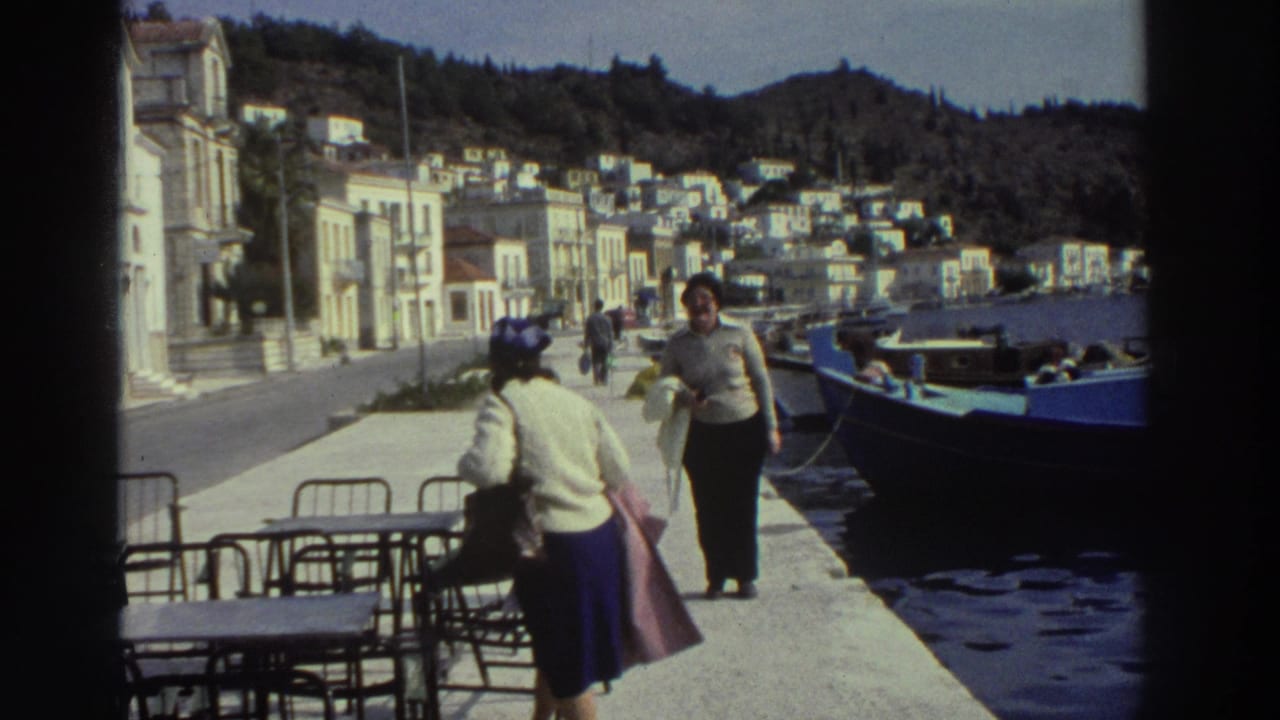 Woman walking past camera on a Greek harbor promenade, 1976; outdoor cafe seating in foreground, blue moored boats and hillside buildings behind.