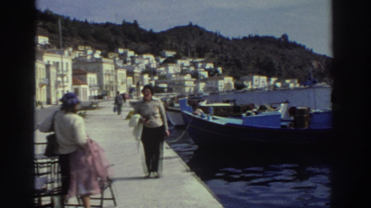 Two women walk along a concrete harbor pier beside a large blue moored fishing boat, white buildings climbing a forested hillside behind.