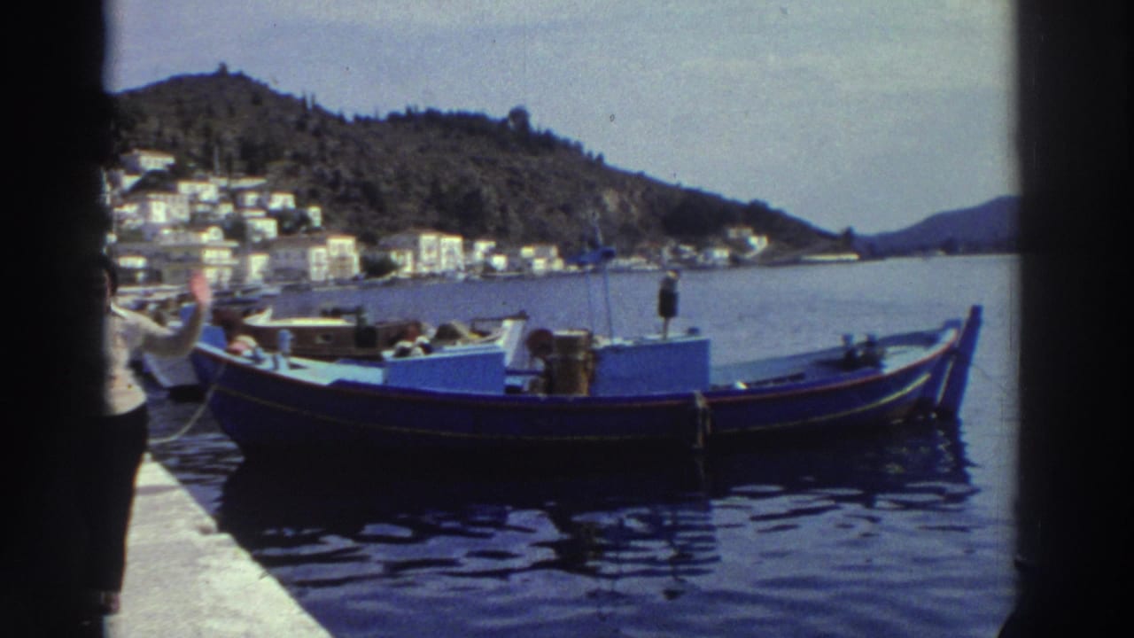 A woman on a concrete pier waves toward the camera beside a blue wooden fishing boat, white hillside buildings and harbor water in the background.