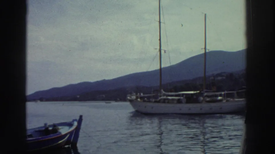 A white two-masted sailing vessel with furled sails crosses what appears to be a Greek harbor circa 1976, a mountain ridgeline and pale overcast sky forming the backdrop.