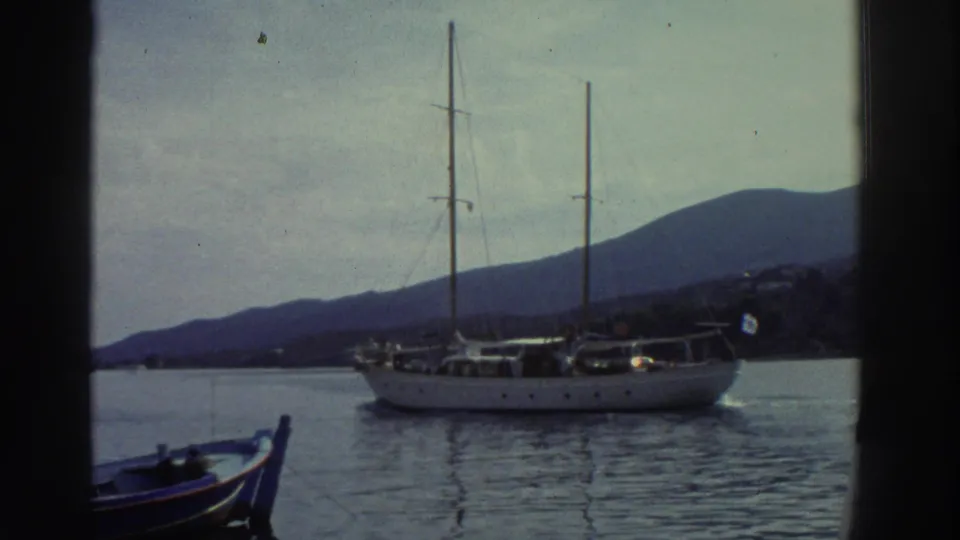 A white two-masted sailing vessel continues across a calm Greek harbor circa 1976, distant mountains and pale sky behind, small blue boat visible at the frame edge.