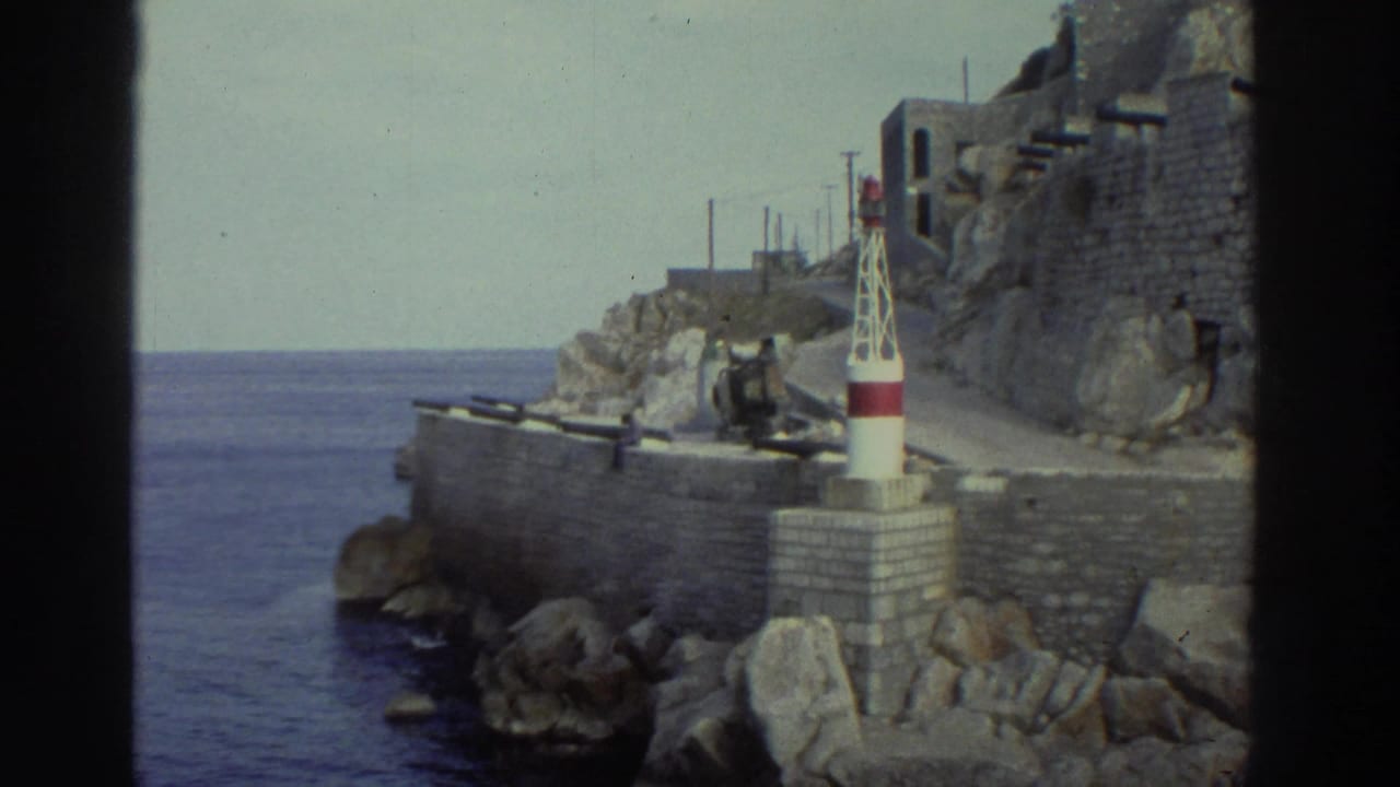 8mm home movie frame, Greece circa 1976 — stone breakwater with a small red-and-white harbor beacon, rocky cliffs, and blue water beyond.
