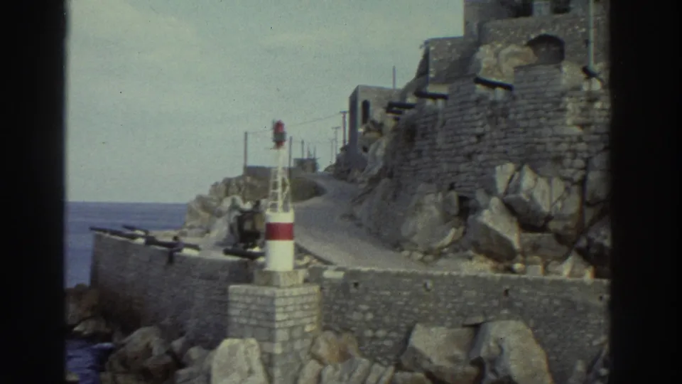 8mm home movie frame, Greece circa 1976 — harbor beacon on stone breakwater, rocky cliffs rising behind, blue sea to the left.
