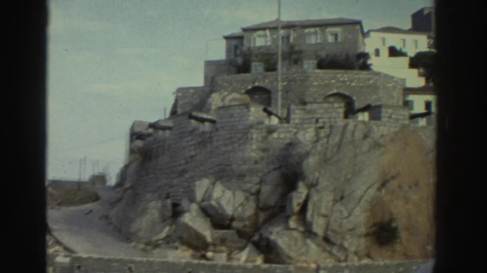 8mm home movie frame, Greece circa 1976 — stone fortress walls with an arched gateway, traditional whitewashed buildings perched on rocky terrain above.