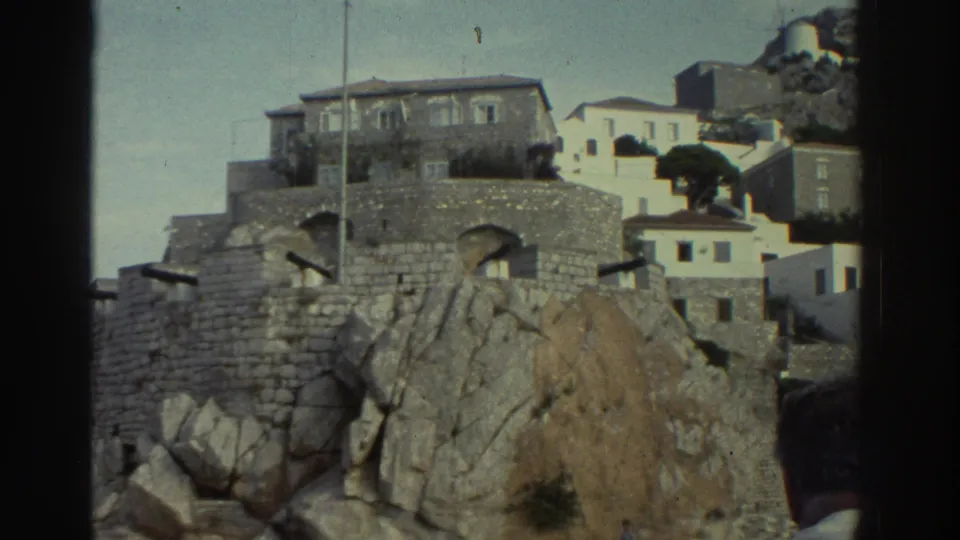 8mm home movie frame, Greece circa 1976 — whitewashed village buildings above stone walls on a rocky coastal hillside, with a tree in the mid-ground.