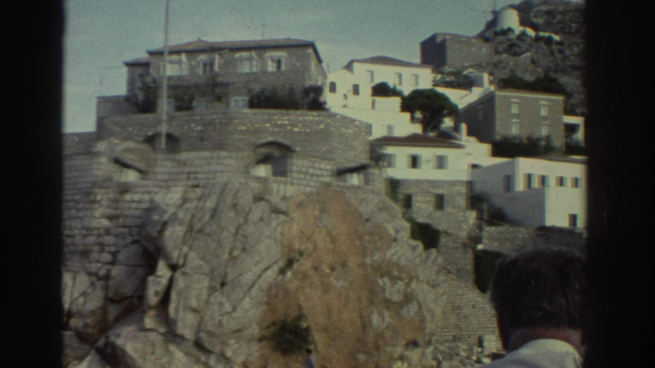 8mm home movie frame, Greece c.1976 — white cubic houses stacked on a rocky coastal hillside with stone walls; back of man's dark hair in lower-right foreground.