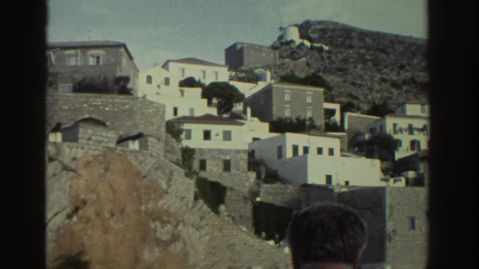 8mm frame, Greece c.1976 — camera pans right across the same hillside cluster of white and stone buildings; back of man's head remains in the lower foreground.