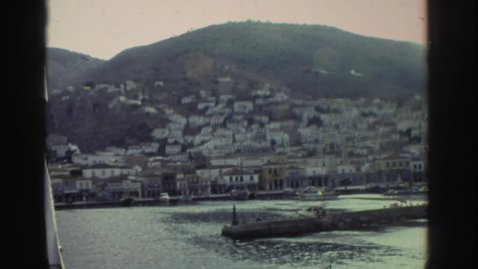 8mm home movie, Greece c.1976 — wide harbour panorama showing white buildings ascending a rounded hillside, waterfront structures, and a breakwater in calm water; boat railing at right edge.