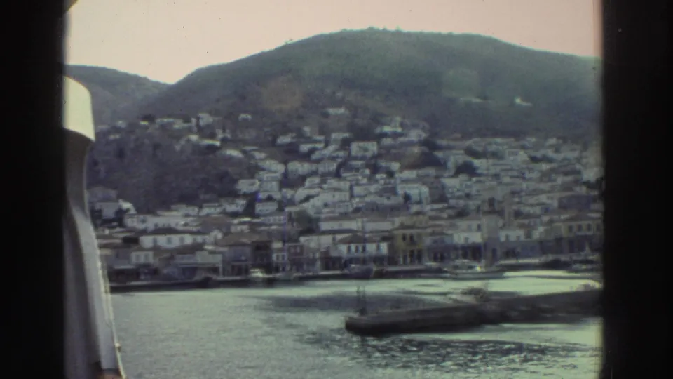 8mm frame, Greece c.1976 — boat's white rail visible at right as camera holds on a hillside harbour town with dense white buildings climbing toward the hilltop and a breakwater in the foreground water.