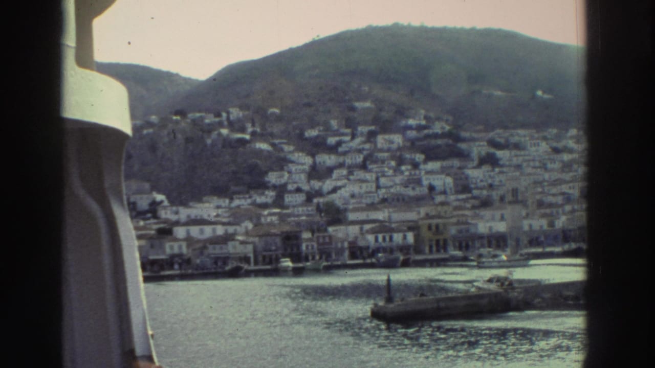 Greek coastal hillside town seen across harbor water from a boat, white buildings cascading down a hillside, stone breakwater visible.