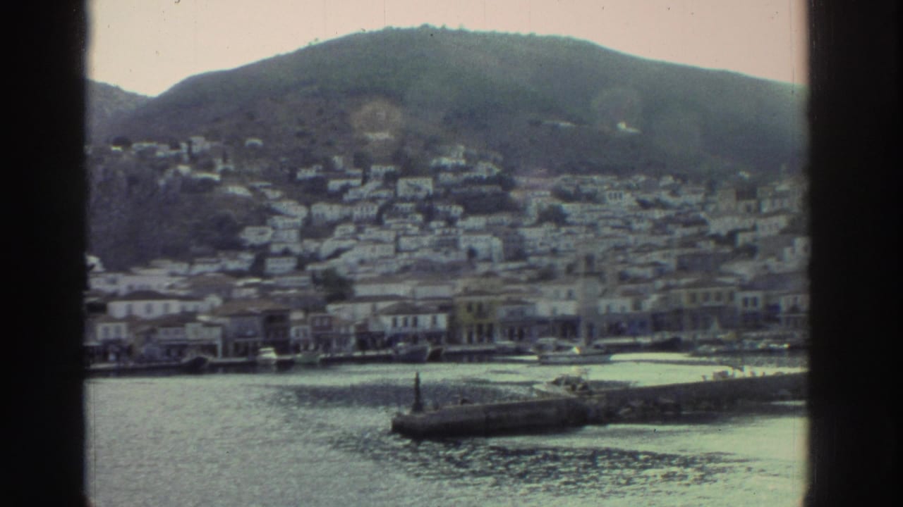 Greek hillside town across calm harbor water from a boat, white buildings on hillside, pier extending into the water.