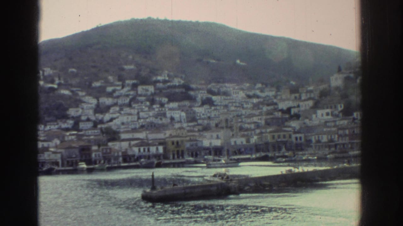 Wider boat-POV of a Greek harbor town, mid-1970s; white and pastel buildings on hillside, waterfront row, stone breakwater, calm water, boat cabin framing.