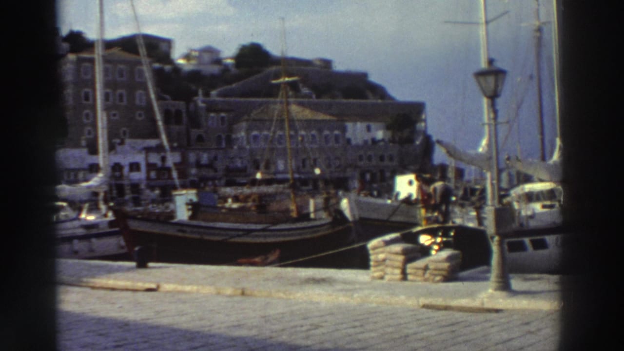 Close view of a Greek harbor quay, mid-1970s; docked wooden sailing boats with tall masts, cobblestone waterfront, classic iron lamppost, stone multi-story buildings behind.
