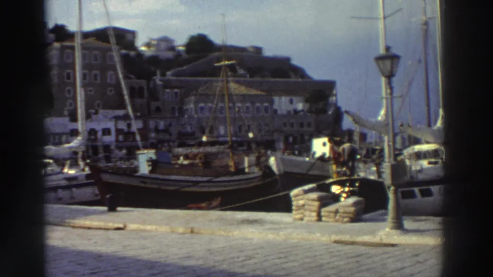 Close view of a Greek harbor quay, mid-1970s; docked wooden sailing boats with tall masts, cobblestone waterfront, classic iron lamppost, stone multi-story buildings behind.