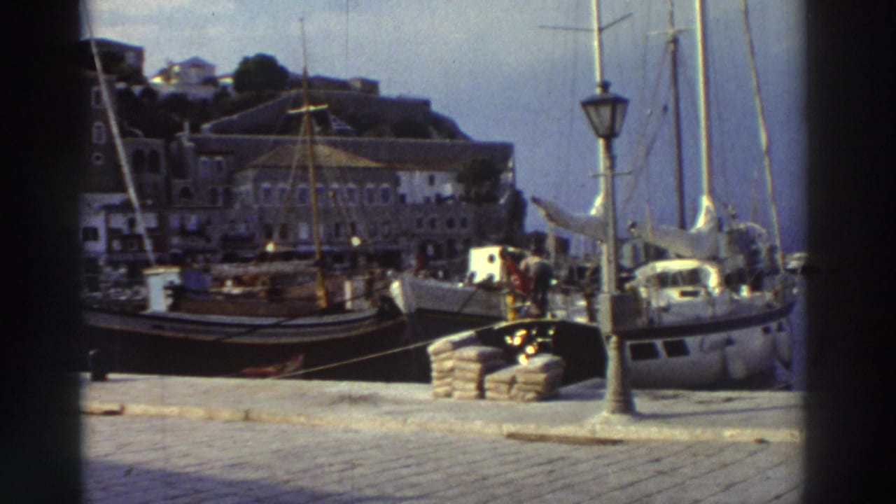 Cobblestone marina quay with docked sailboats, tall masts, and an ornate lamppost; hillside town with stone and white buildings rises in the background.