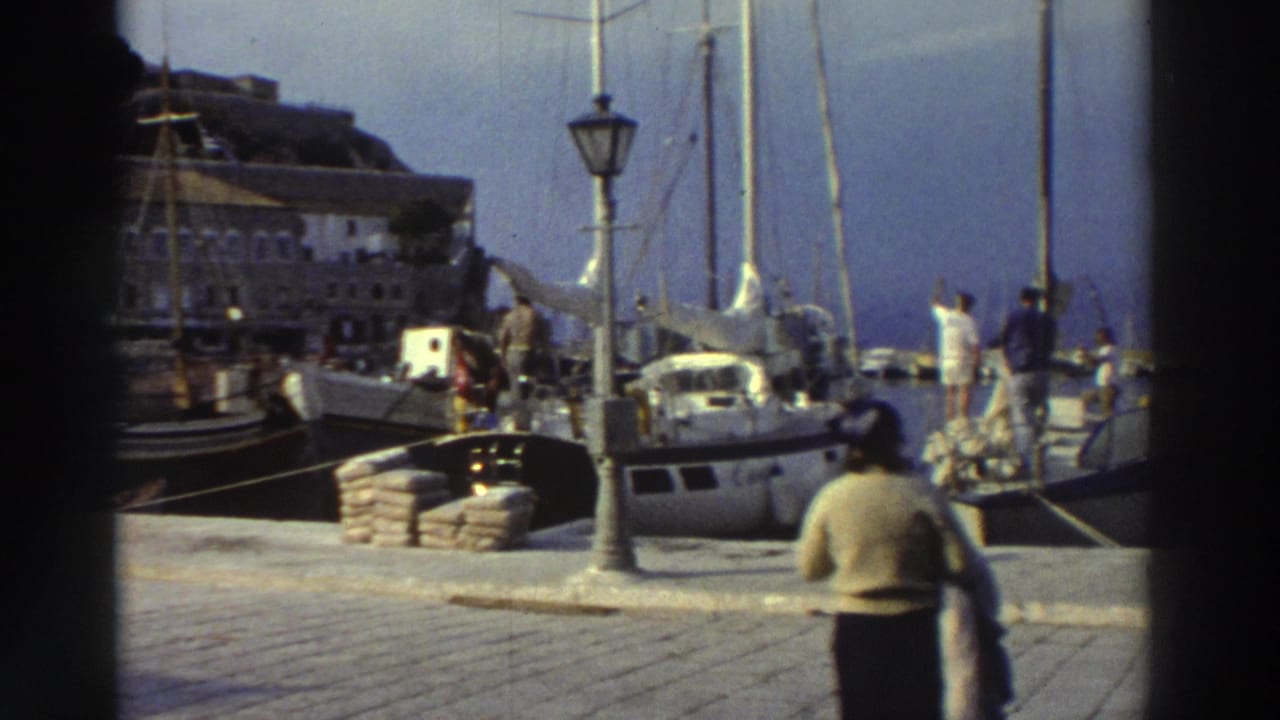 Marina quay with docked sailboats and lamppost; a figure in a light top and dark trousers walks away from camera in the right foreground.