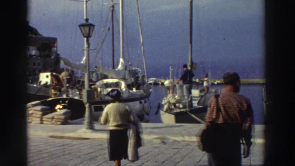 8mm frame from a Greek marina, circa 1976, showing a woman and man walking along the cobblestone quay with docked sailboats, an ornate lamppost, and a hillside town behind them.