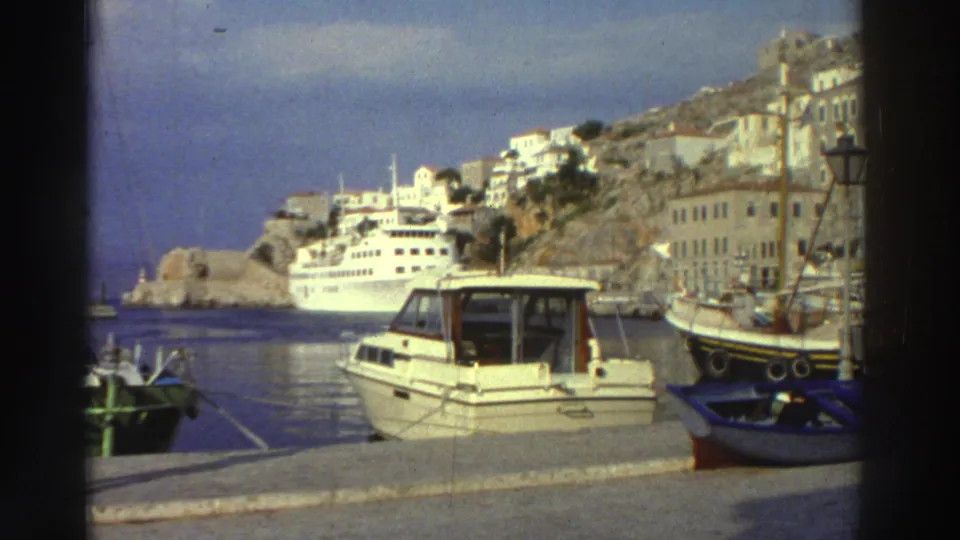 8mm footage of a Greek island harbor, circa 1976; a large white cruise ship anchors offshore while a motorboat and small vessels occupy the foreground, and a steep hillside town with white-washed buildings fills the background.