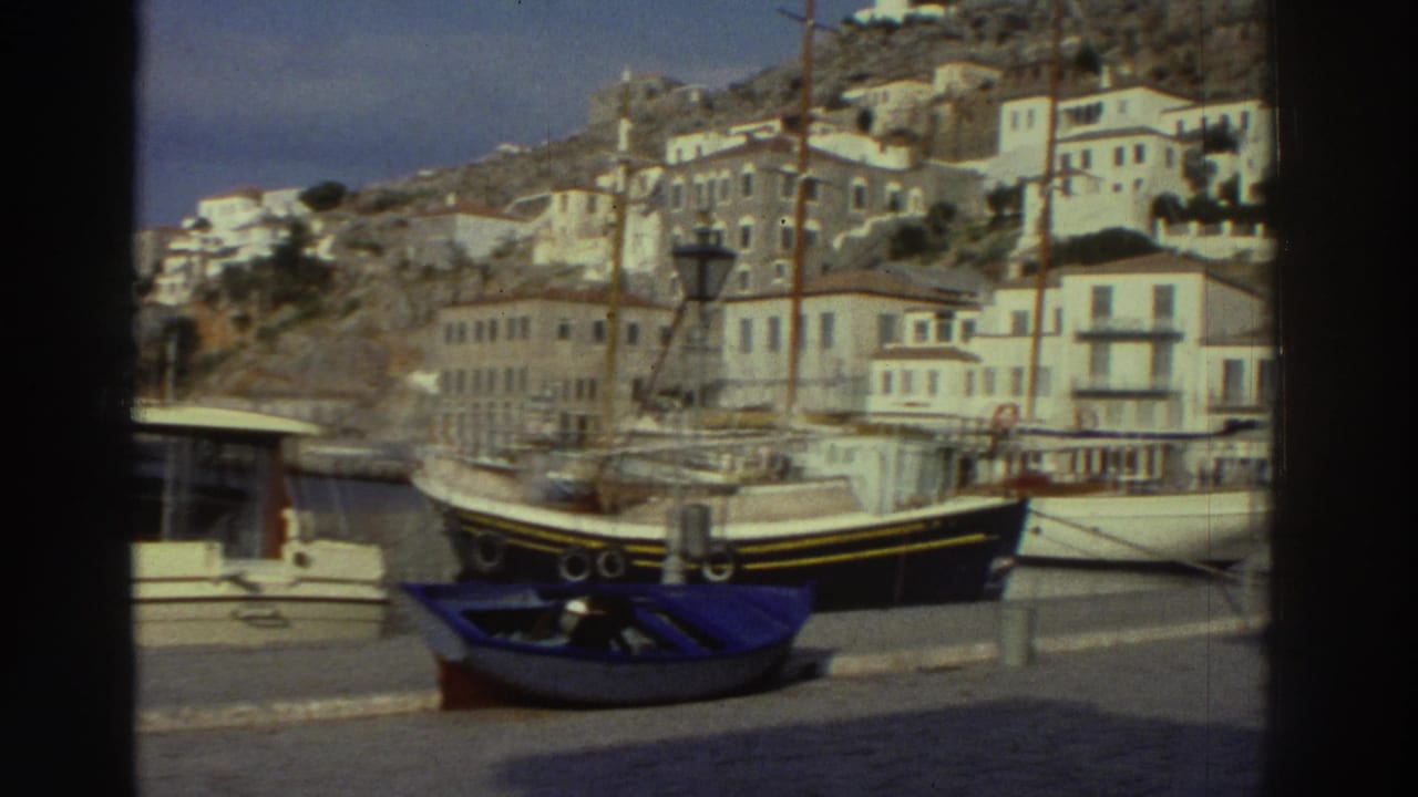 Continued harbor view showing white motorboat and dark-hulled fishing vessel at the quay, white buildings on hillside, cruise ship in the far background.