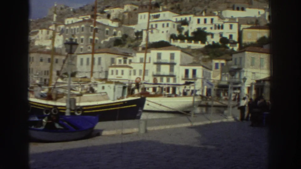 8mm home movie of a Greek harbor waterfront, 1976: fishing boat moored at quay, people strolling the promenade, white multi-story buildings with ground-floor shops rising behind.