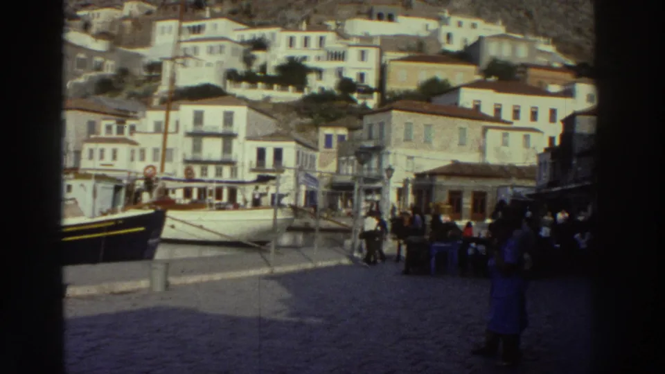 Final frame of 8mm Greek harbor footage, 1976: fishing boat at quay, cluster of pedestrians on the waterfront, tiered white buildings with ground-floor shops on the hillside.