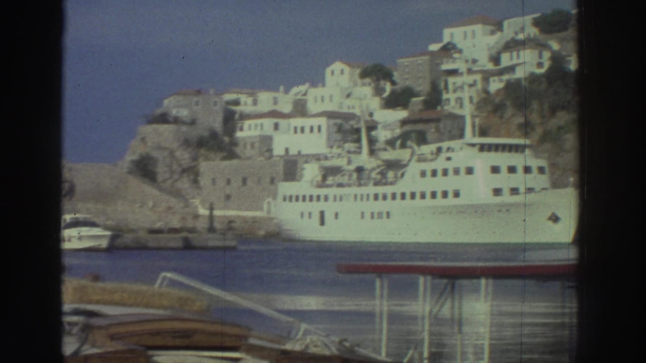 White passenger ferry docked in a Greek harbor, wooden fishing boats in foreground, white buildings on rocky hillside behind.