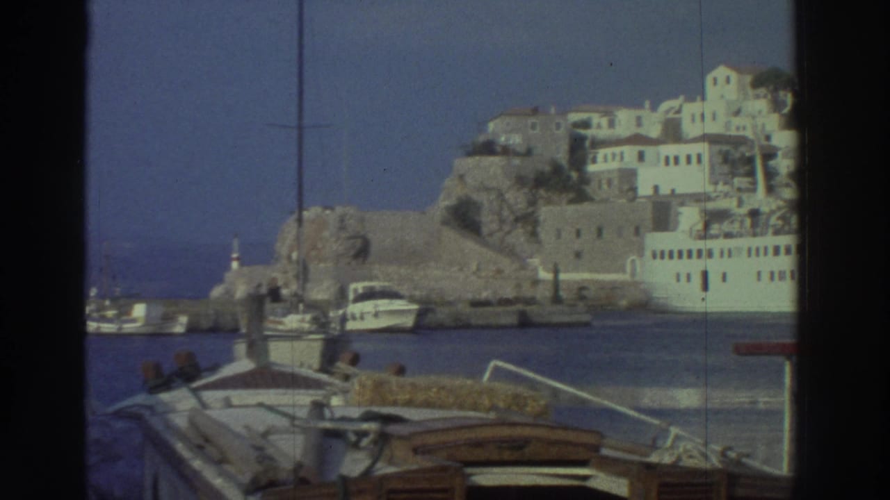 Pan across Greek harbor; rocky hillside with white cubic buildings on right, small vessels moored in foreground.