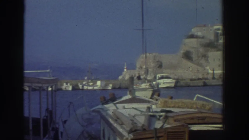 Cluster of small moored vessels at a Greek island quay in 1976, with a rocky headland and harbor entrance in the background.