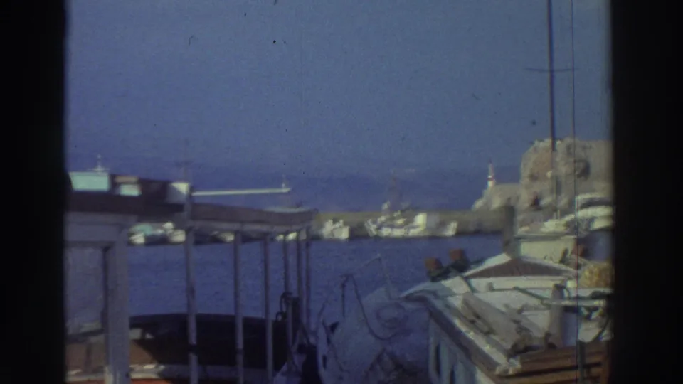 Greek island harbor at pan endpoint in 1976, showing clustered small vessels, open water, and a distant coastline.