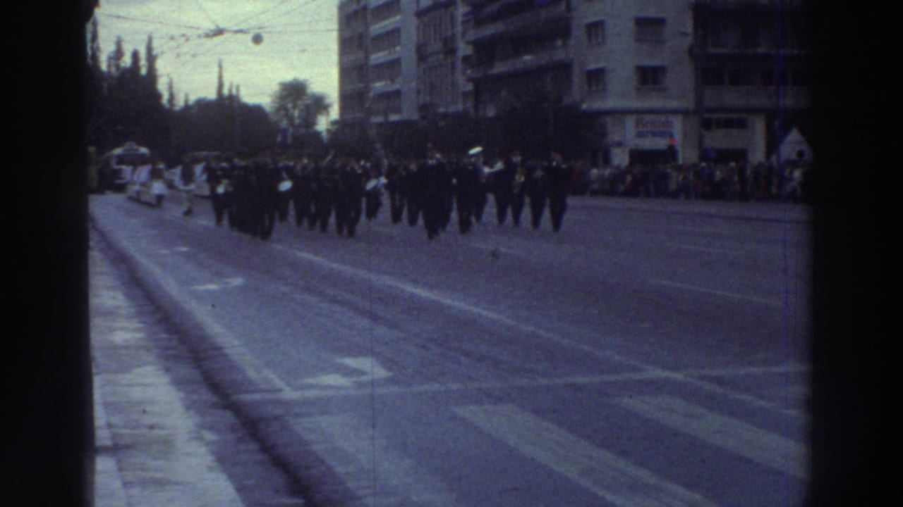 Wide-angle view of a military marching column on a broad Greek city boulevard, 1976; spectators line the pavement with mid-century apartment buildings behind.