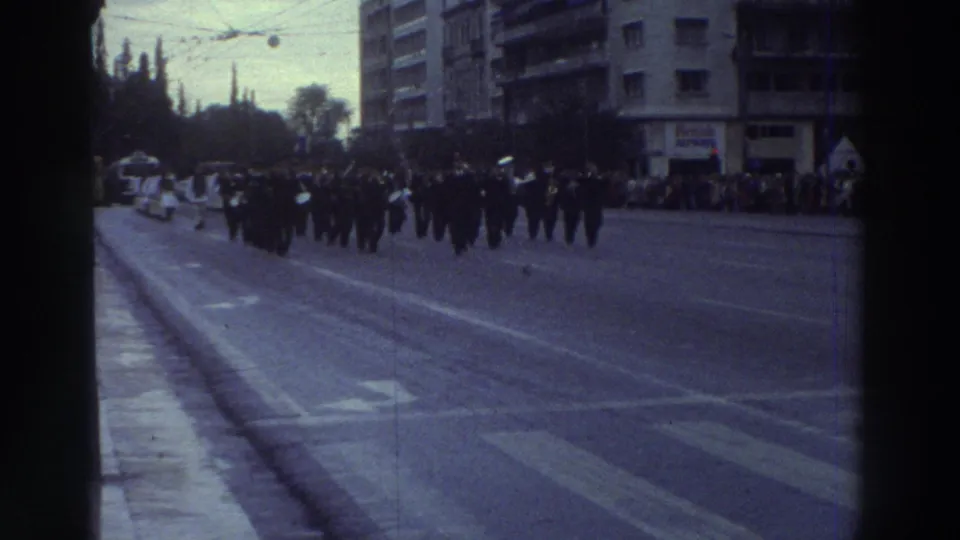 Wide-angle view of a military marching column on a broad Greek city boulevard, 1976; spectators line the pavement with mid-century apartment buildings behind.