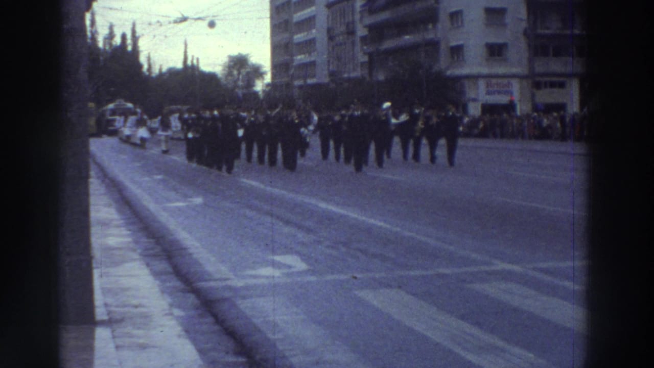 Wide shot of uniformed military column continuing march on broad boulevard; spectators and apartment blocks visible on both sides.
