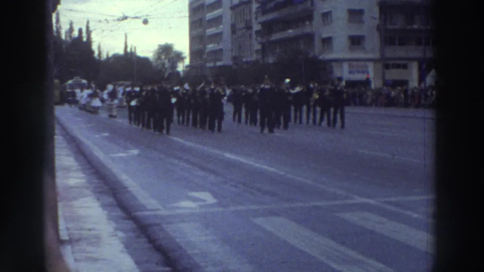 Greek city parade route, 1976; tram tracks, trolleybus catenary wires, and a prominent modernist apartment building frame the marching military column.