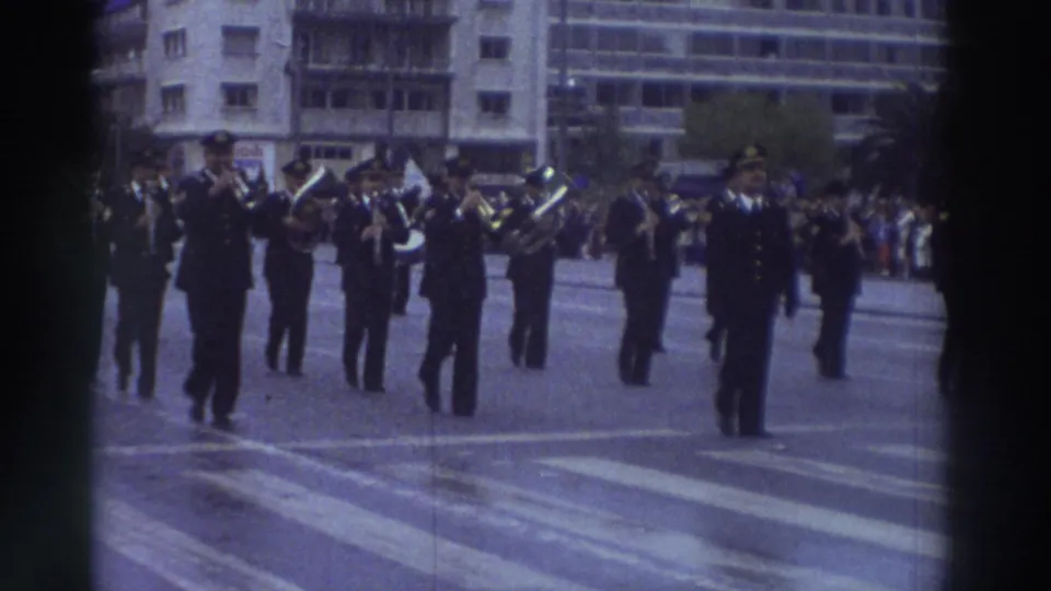 Close view of a Greek military brass band marching in a 1976 parade; musicians in dark dress uniforms play brass instruments with a crowd watching at right.