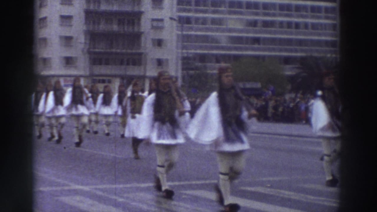 Close view of Evzone-style ceremonial marchers in white pleated skirts and dark vests, filmed on 8mm in Greece circa 1976, with a large modern building in the background.
