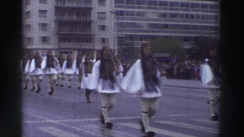 Close view of Evzone-style ceremonial marchers in white pleated skirts and dark vests, filmed on 8mm in Greece circa 1976, with a large modern building in the background.