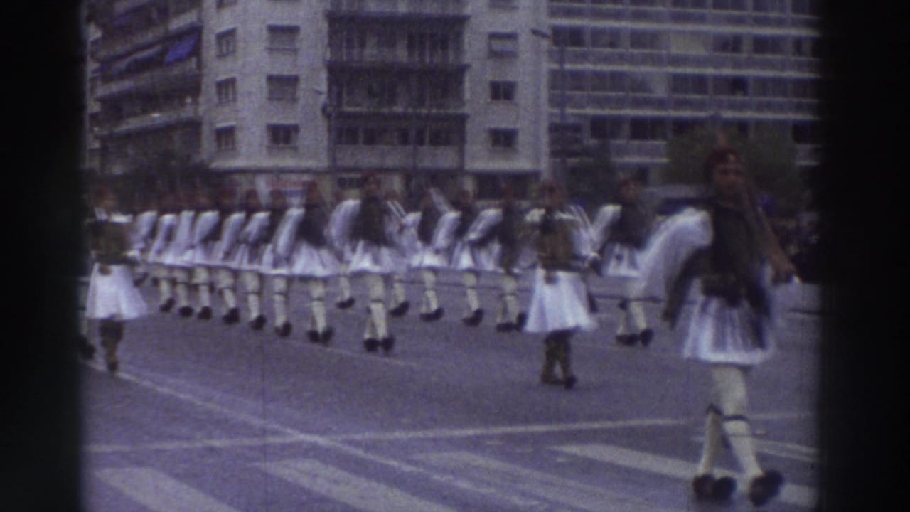 Wide column of men in white traditional costumes marching in unison, a large building visible at upper right.