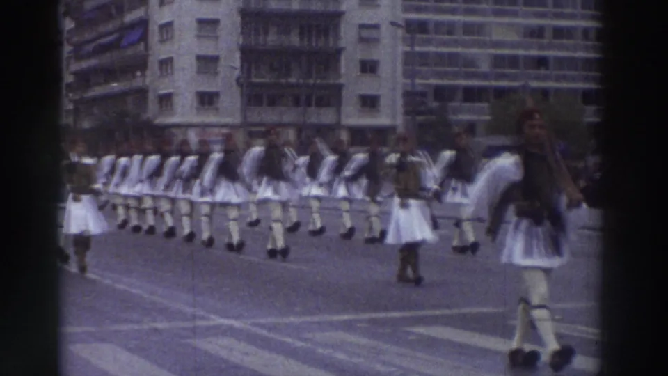 Extended column of Evzone-style ceremonial marchers in white foustanella stretching across a wide open area, filmed on 8mm in Greece circa 1976.