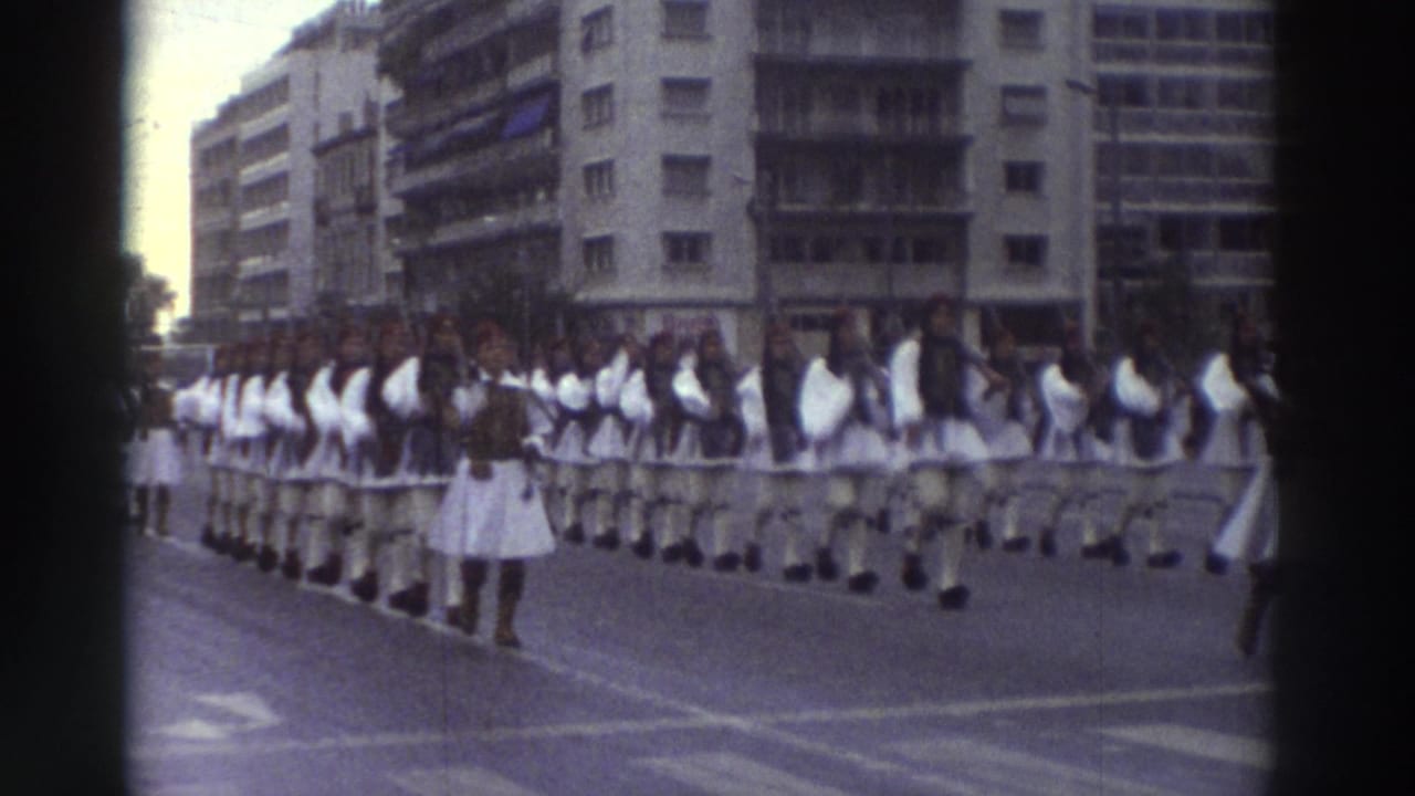 Ceremonial parade column in white traditional dress moving along a wide Greek city boulevard, circa 1976, with mid-century buildings and overhead tram wires framing the march.
