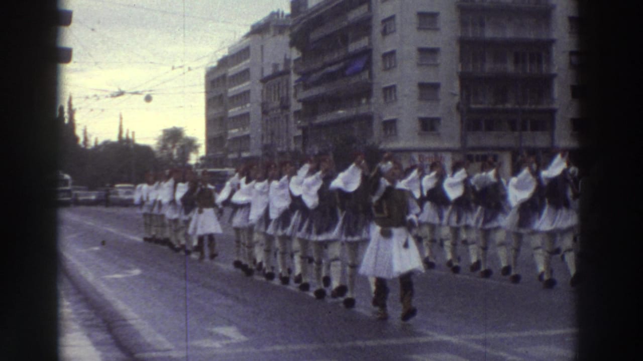 Long ceremonial parade column in white Evzone-style dress on a wide Greek city boulevard, circa 1976, with multi-story buildings and overhead wires framing the procession.