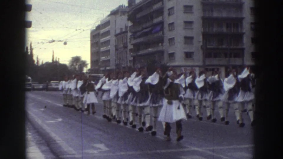 Long ceremonial parade column in white Evzone-style dress on a wide Greek city boulevard, circa 1976, with multi-story buildings and overhead wires framing the procession.