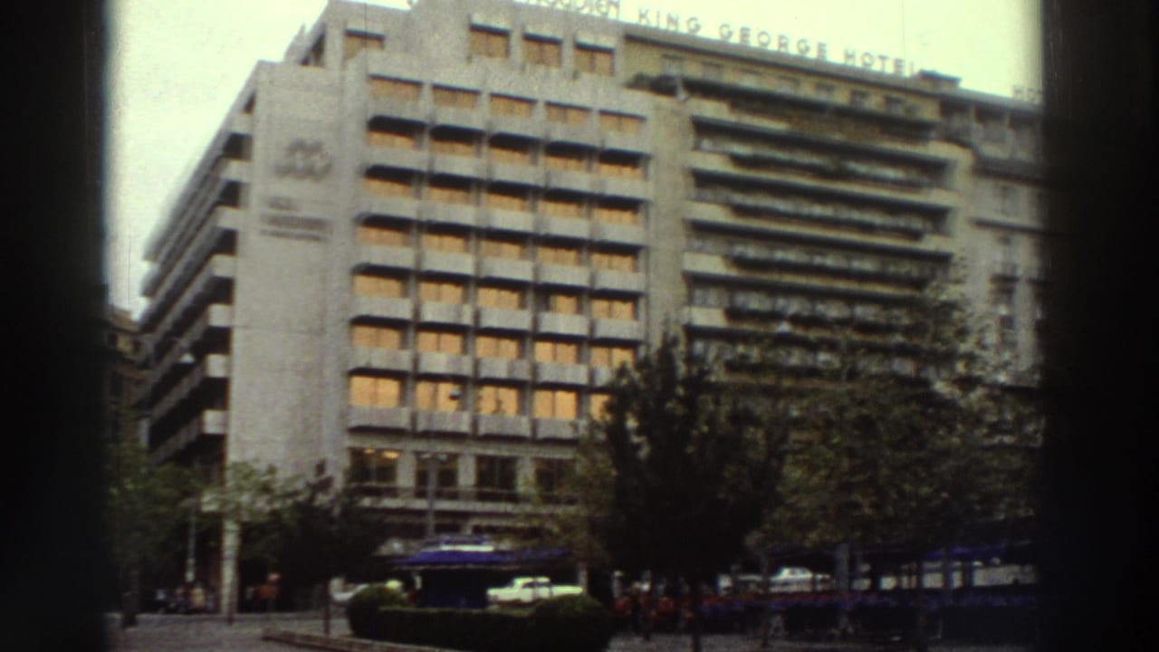 Large multi-story hotel with horizontal banded facade and readable roofline signage, blue café umbrellas and street trees in foreground.