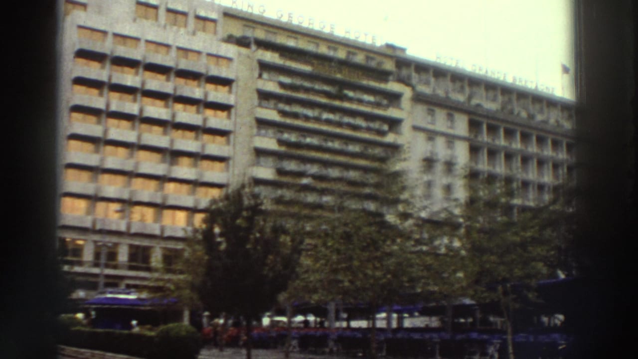 Continued pan of the same large hotel building showing roofline lettering, rows of windows, and blue umbrellas below street trees.