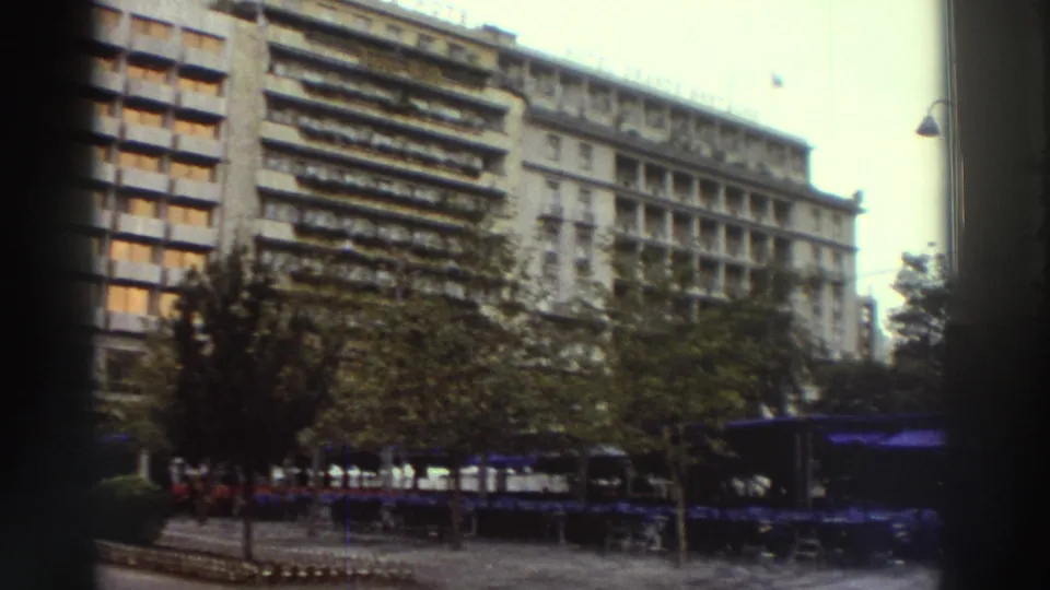 Angled view of the hotel in Athens, Greece, circa 1976, showing the multi-story modernist facade, ground-level outdoor seating with blue umbrellas, and mature trees.