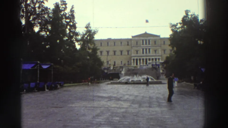Wide view of what appears to be a central Athens plaza circa 1976, showing a fountain, a solitary pedestrian, outdoor café umbrellas, trees, and a neoclassical civic building in the background.