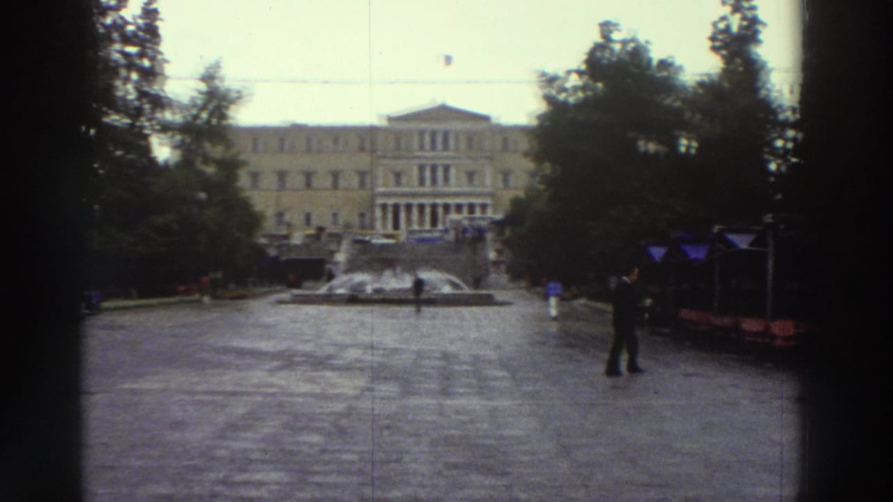 Super 8 wide shot of a wet Greek city square circa 1976, with a neoclassical public building, blue market awnings, a central fountain, and a lone pedestrian crossing the plaza.