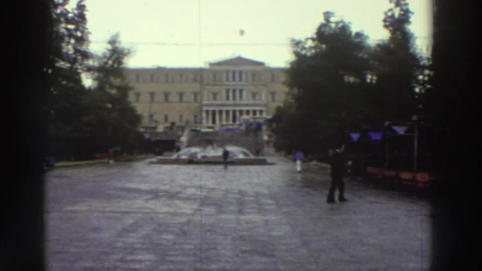 Super 8 wide shot of a wet Greek city square circa 1976, with a neoclassical public building, blue market awnings, a central fountain, and a lone pedestrian crossing the plaza.