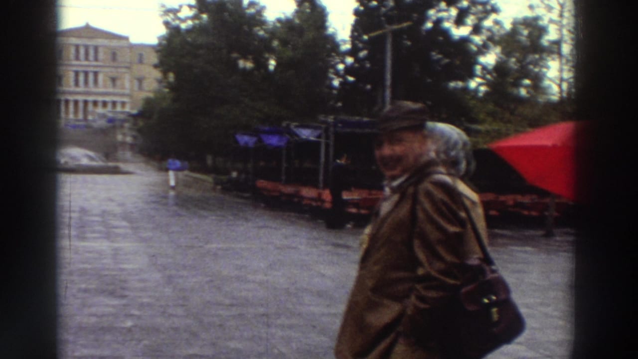 Man in a brown coat and dark cap turns to smile at the camera in a city square, with a red umbrella visible at the edge of the frame.